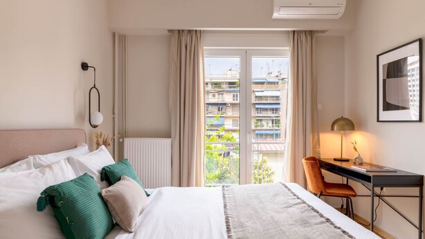 A hotel room with a bed, a desk, a chair, a lamp, and a view of buildings through a window. - Charismatic 2BR Apartment in Hilton (Athina)