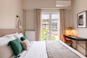 A hotel room with a bed, a desk, a chair, a lamp, and a view of buildings through a window.
