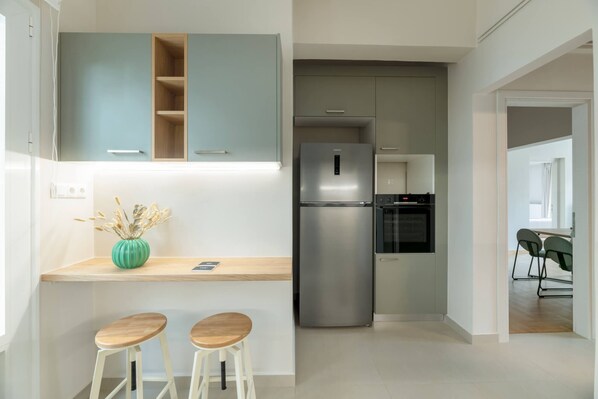 A modern kitchen with a stainless steel refrigerator, a built-in oven, and a wooden countertop. There are two wooden stools and a green vase with dried plants.