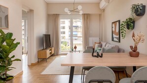 A modern living room with a wooden dining table, grey chairs, a sofa, and a TV on a wooden stand. There are potted plants and a framed picture on the walls. - Charismatic 2BR Apartment in Hilton (Athina)