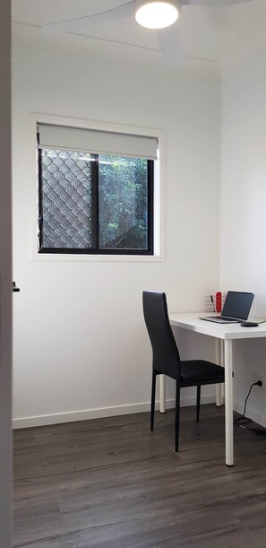 Une pièce avec un bureau blanc, une chaise noire, un ordinateur portable et une fenêtre munie d’un moustiquaire métallique.
