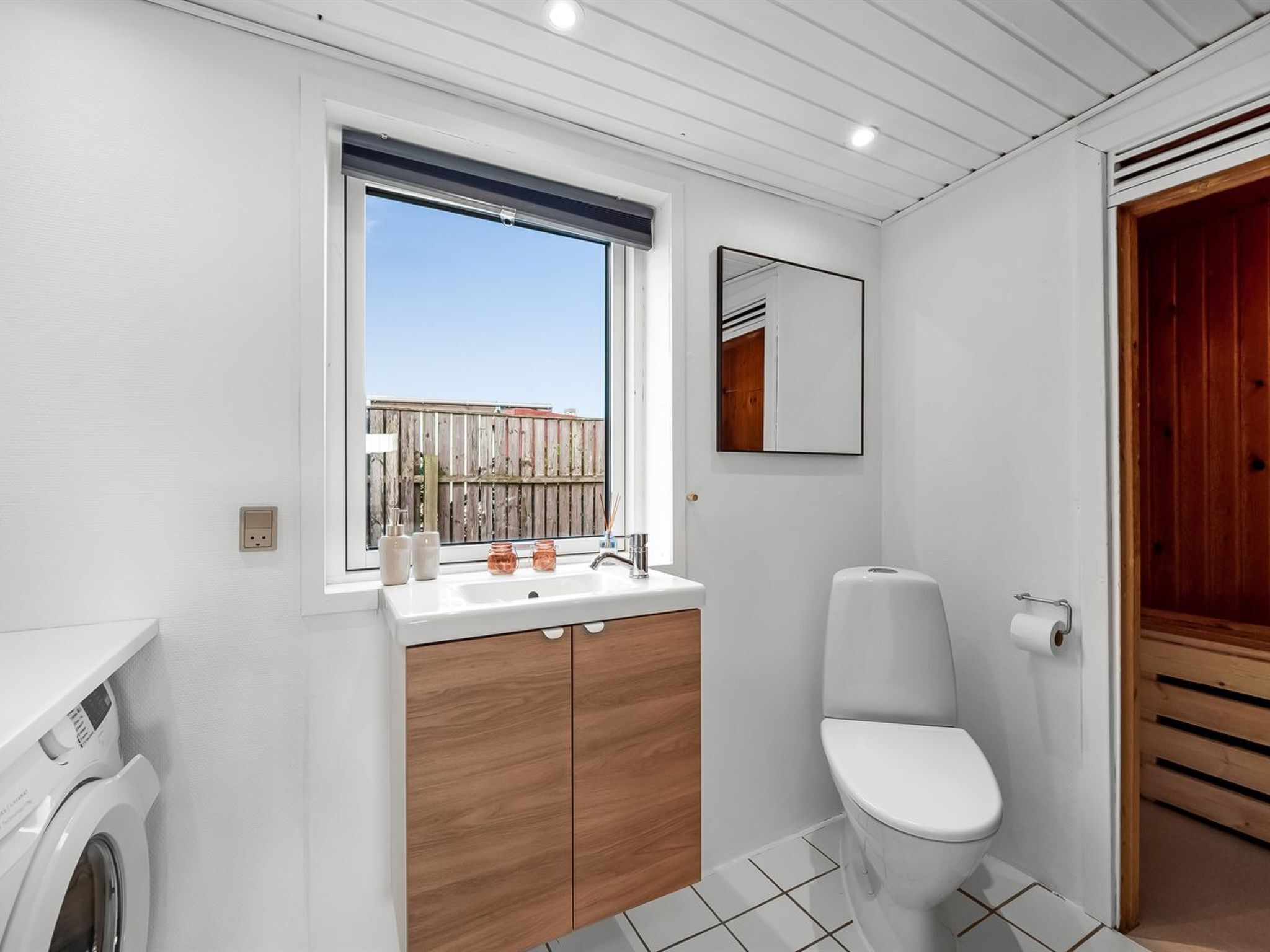 A compact, modern bathroom with a window, a sink, a toilet, and a wooden cabinet.
