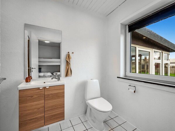 Un baño con un lavamanos blanco, un mueble de madera y un inodoro. Hay una ventana con vistas a una casa.