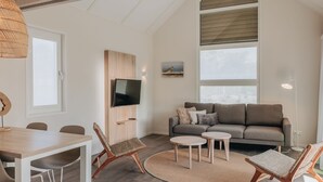 A modern living room with a wooden dining table, two chairs, a grey sofa, and a TV mounted on a wooden panel. - Lodge in Zeeland Near Nieuwvliet Beach (Nieuwvliet)