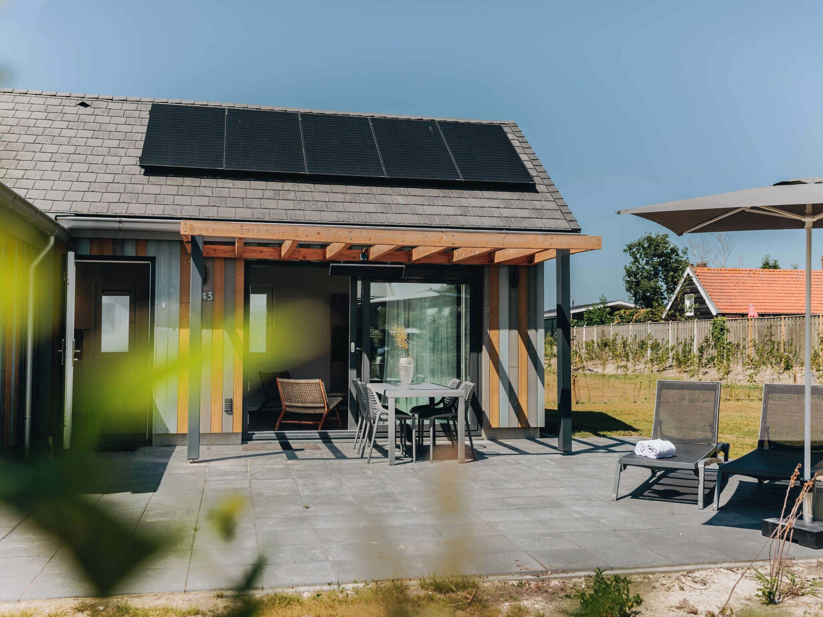 Une maison moderne dotée d’un toit équipé de panneaux solaires, d’un patio couvert avec des meubles d’extérieur, et offrant une vue dégagée sur les environs.