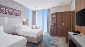 A hotel room with two beds, a desk with a phone and remote, a TV, a large window, and a door with a mirror. - Nur Al Naseem  (Makkah)