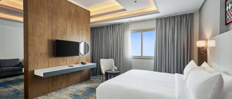 Une chambre d’hôtel avec un grand lit, un téléviseur à écran plat fixé sur un panneau en bois, un bureau avec une lampe, une chaise et une vue sur le ciel par la fenêtre.