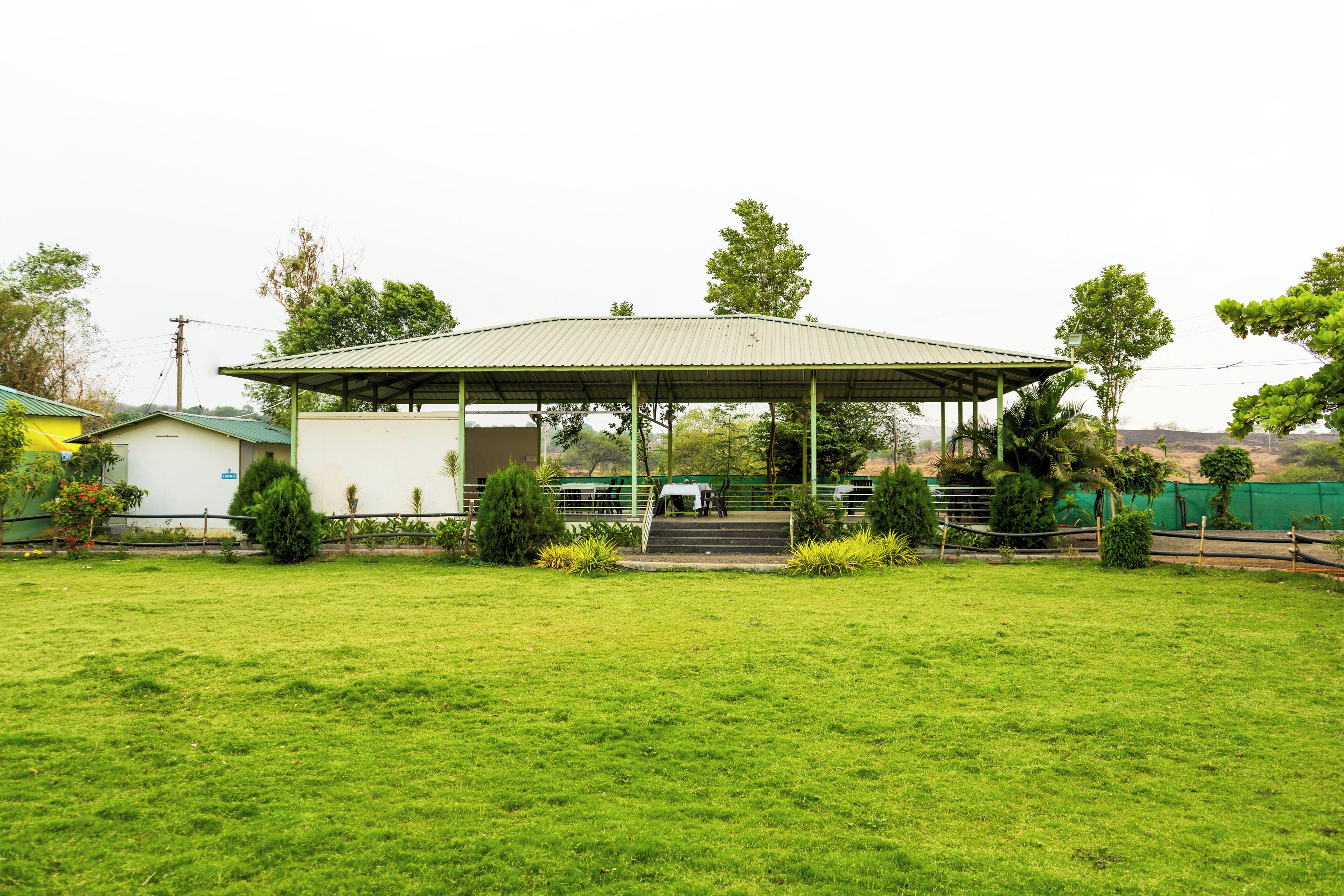 A covered pavilion with a roof, surrounded by a grassy field and a few trees.