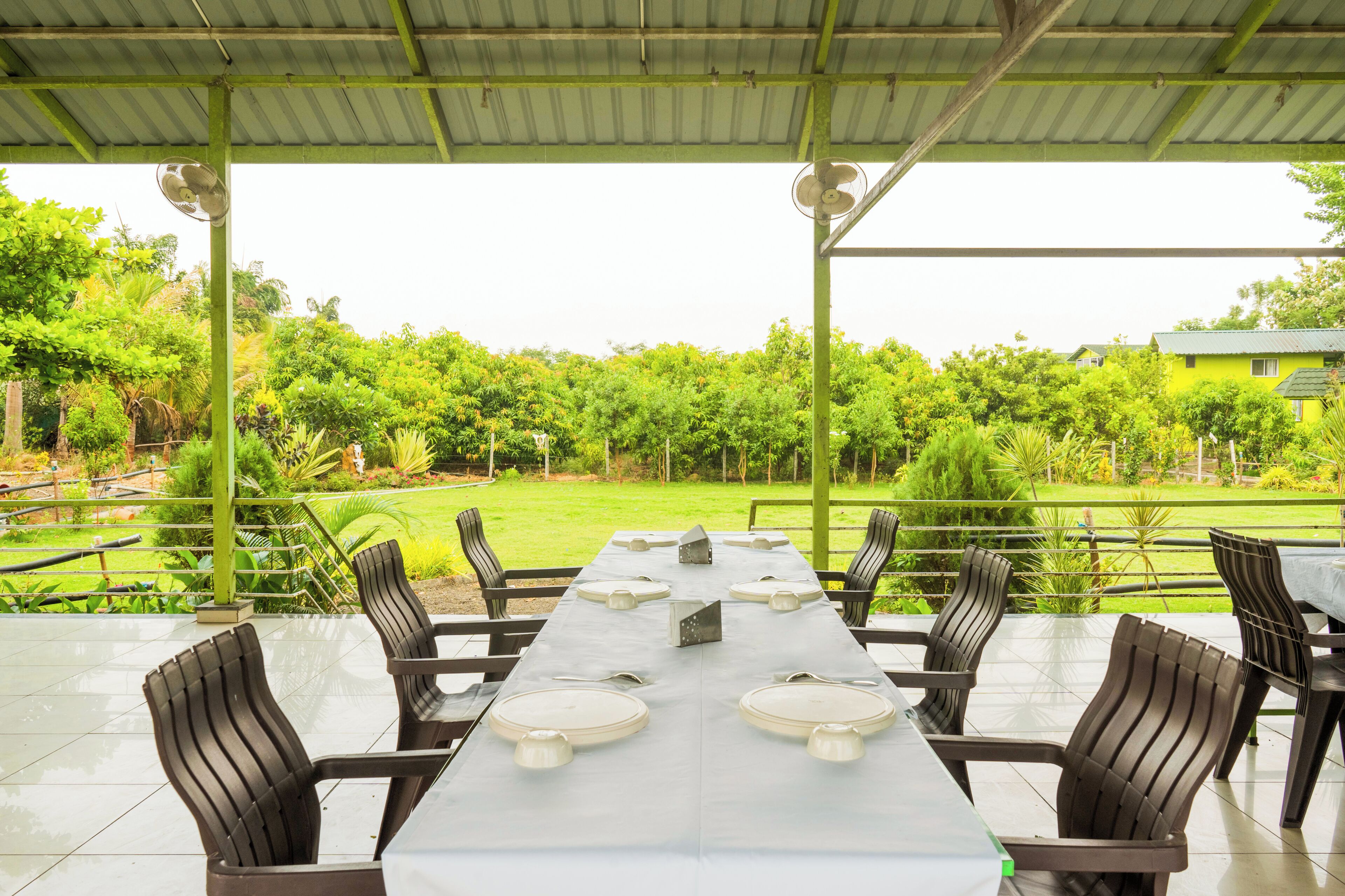 Un espace extérieur abrité, aménagé pour les repas, avec une longue table dressée pour six personnes, entourée de chaises, et offrant une vue sur un jardin luxuriant et un bâtiment en arrière-plan.