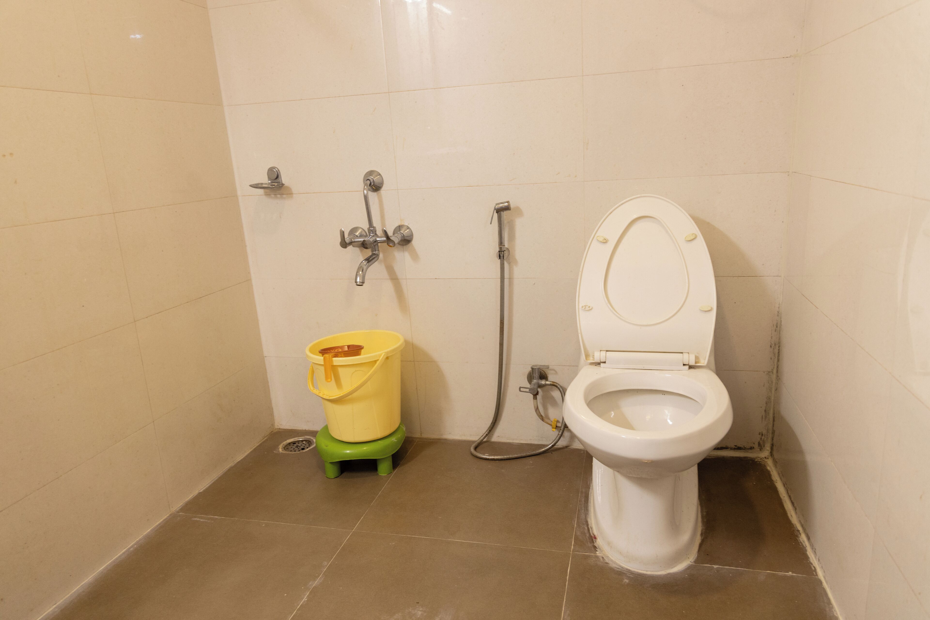 Une petite salle de bain propre, avec des toilettes, une douche et un seau jaune posé sur un tabouret vert.