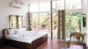 A bedroom with a large bed, a ceiling fan, an air conditioning unit, and a view of a garden through large windows. - The Riverine Stays by Igatpuri (Igatpuri)