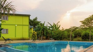 A swimming pool with a green building in the background, surrounded by lush greenery. - The Riverine Stays by Igatpuri (Igatpuri)