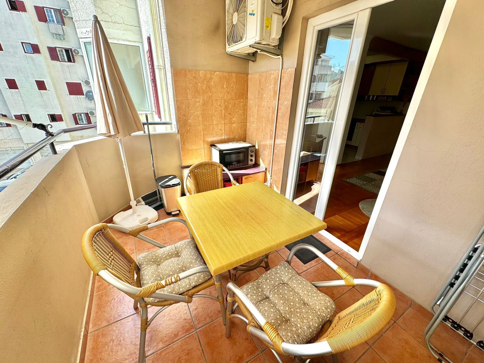 Un balcón con una mesa amarilla, sillas de mimbre, una kitchenette pequeña y vista a un edificio con contraventanas rojas.