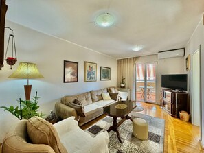 Una sala de estar con un sofá, una mesa de centro, un televisor y un balcón con mobiliario exterior.