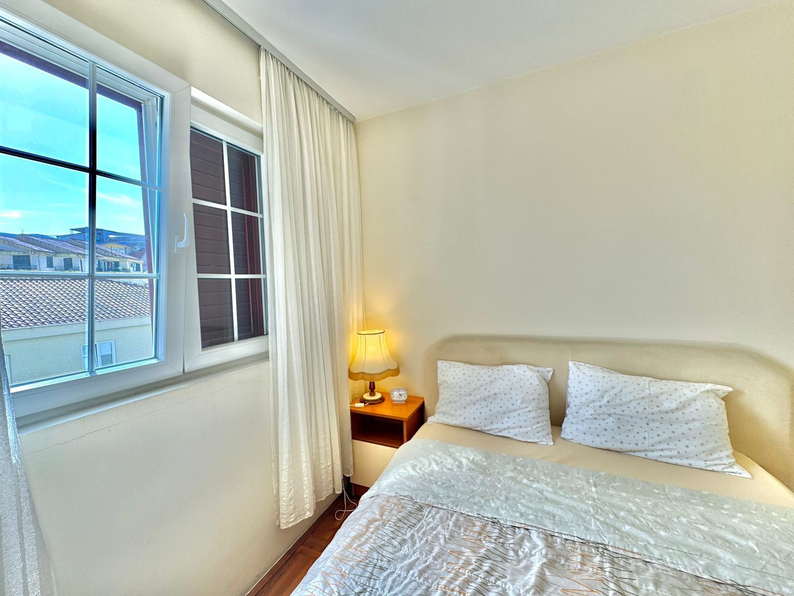 A hotel room with a bed, a nightstand with a lamp, a window with curtains, and a view of buildings outside.