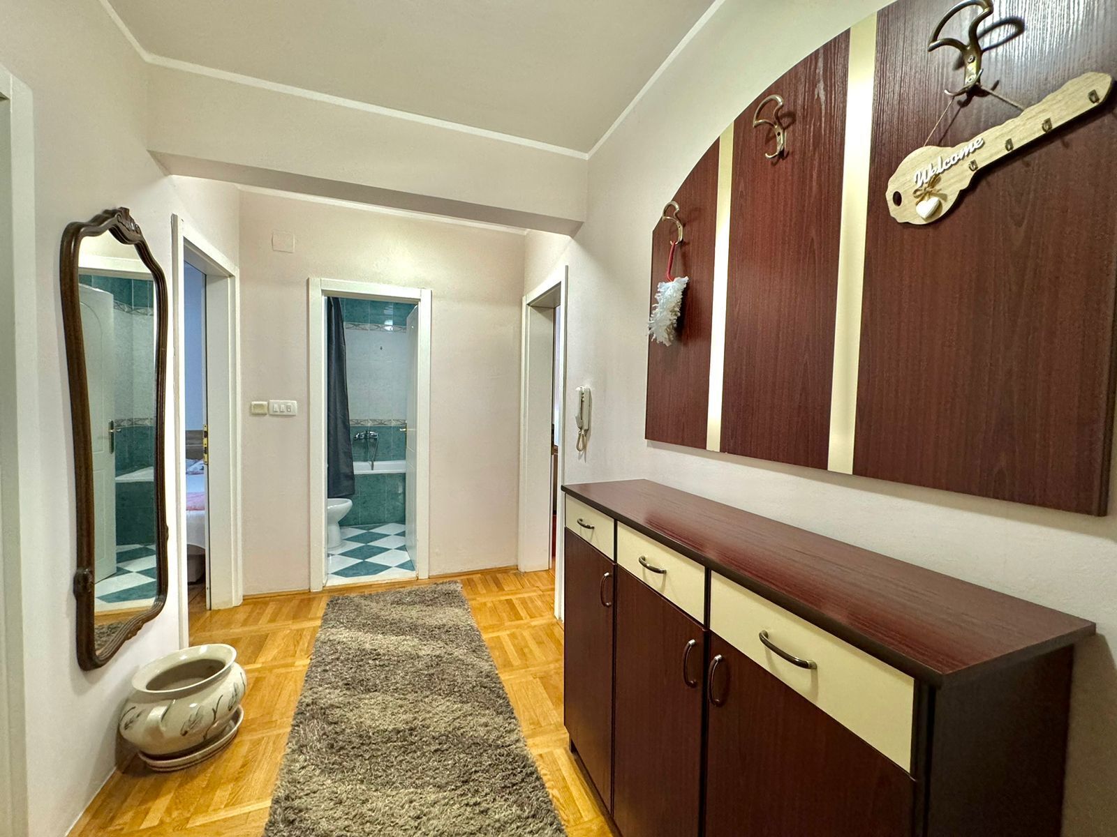 A hallway with a wooden floor, a rug, a cabinet with drawers, a mirror, and a door leading to a bathroom.
