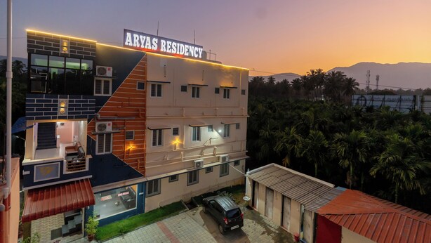 Un immeuble à plusieurs étages, appelé « Aryas Residency », présente un design moderne, avec une façade combinant des éléments en brique et en crépi, et une voiture garée dans l’allée.