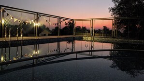 A swimming pool with a metal fence and string lights, surrounded by trees and a sunset in the background. - Hotel Baga Stays By Coral (Calangute)