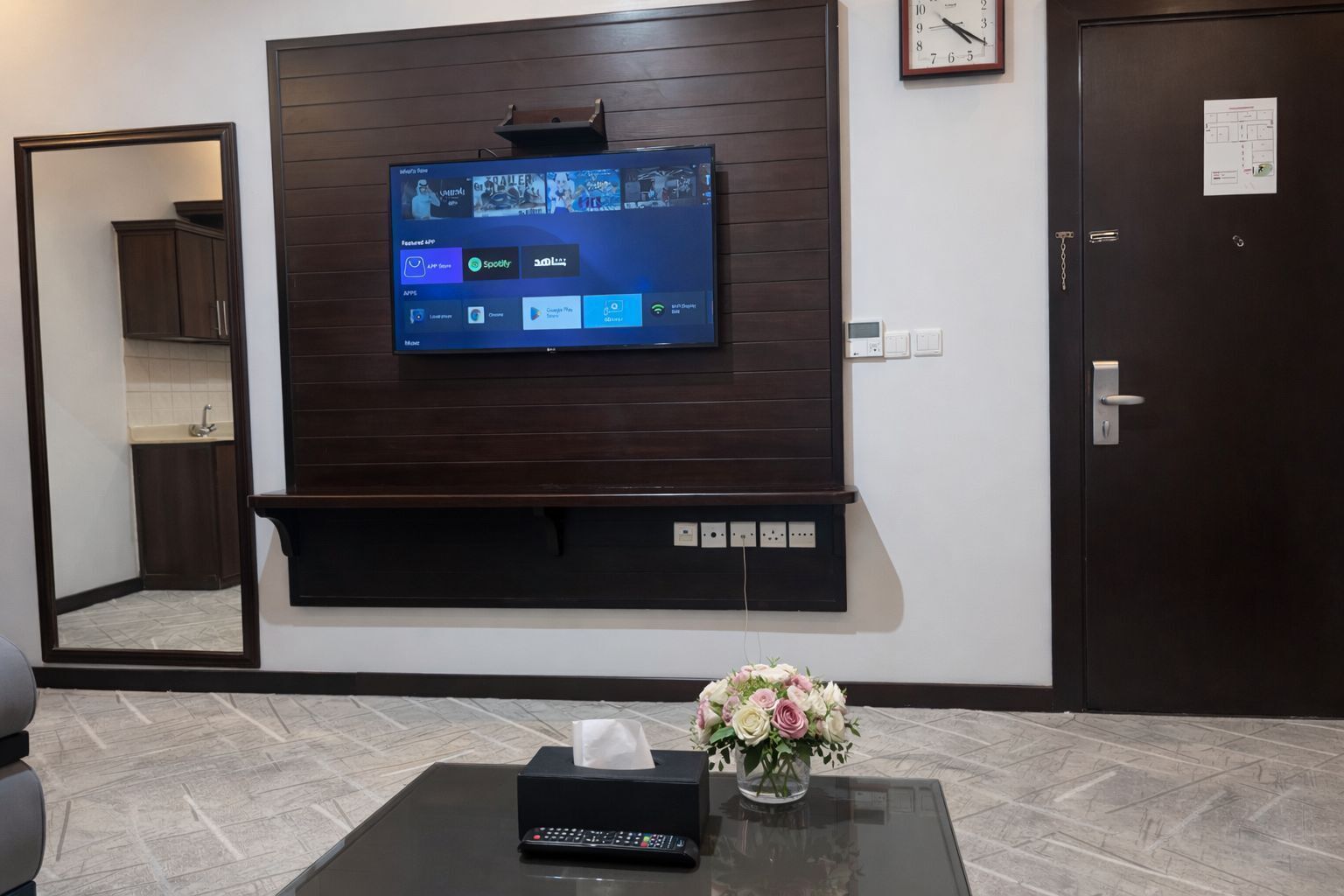 A hotel lobby with a mounted TV displaying various streaming services, a reception desk with a tissue box and vase of flowers, and a clock on the wall.