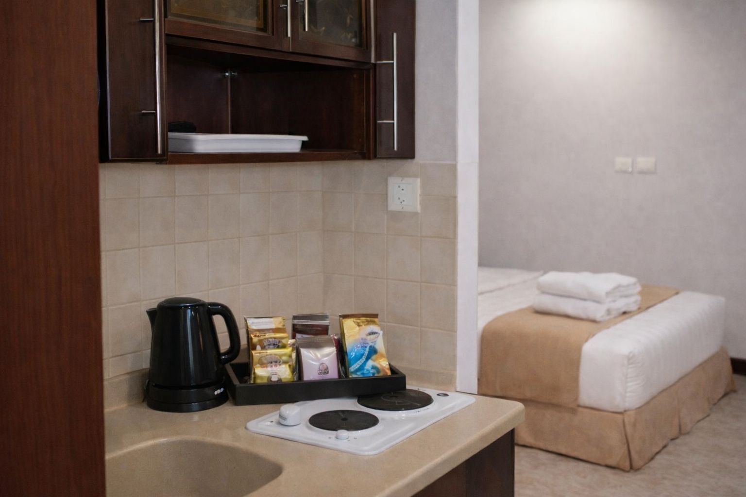 A kitchenette with a coffee maker, tea bags, and a small stove. There is a bed with a beige mattress and white linens.