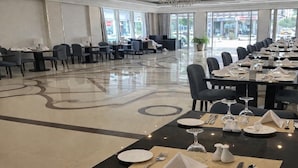 Un comedor de hotel con mesas dispuestas para el servicio, una araña y vistas de la calle a través de amplias ventanas.