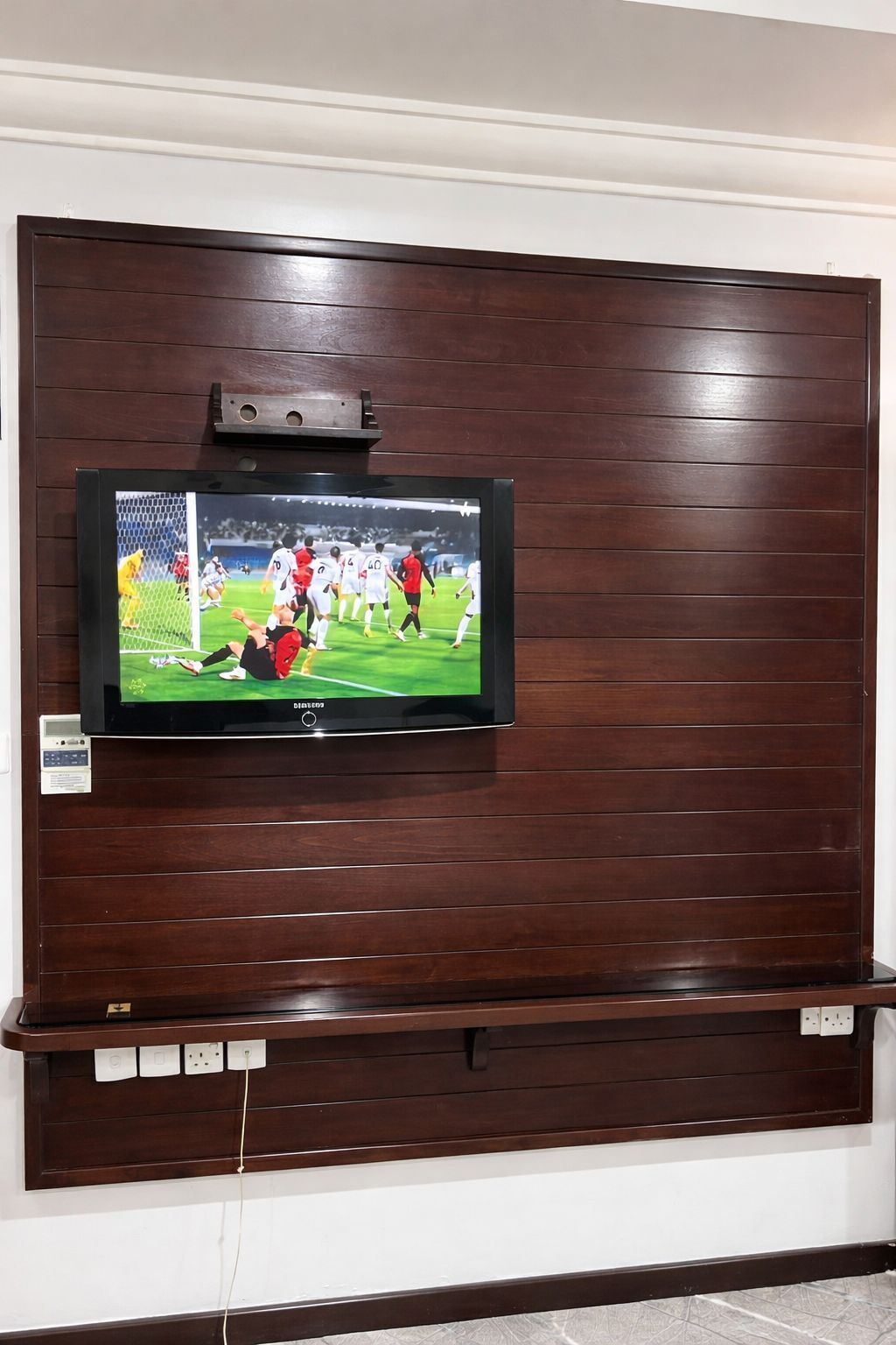 Televisão de tela plana fixada em parede de madeira, com uma prateleira acima.