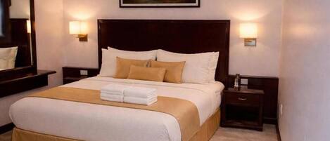Habitación de hotel con una cama grande, dos mesitas de noche, un tocador, un cuadro en la pared y un espejo.