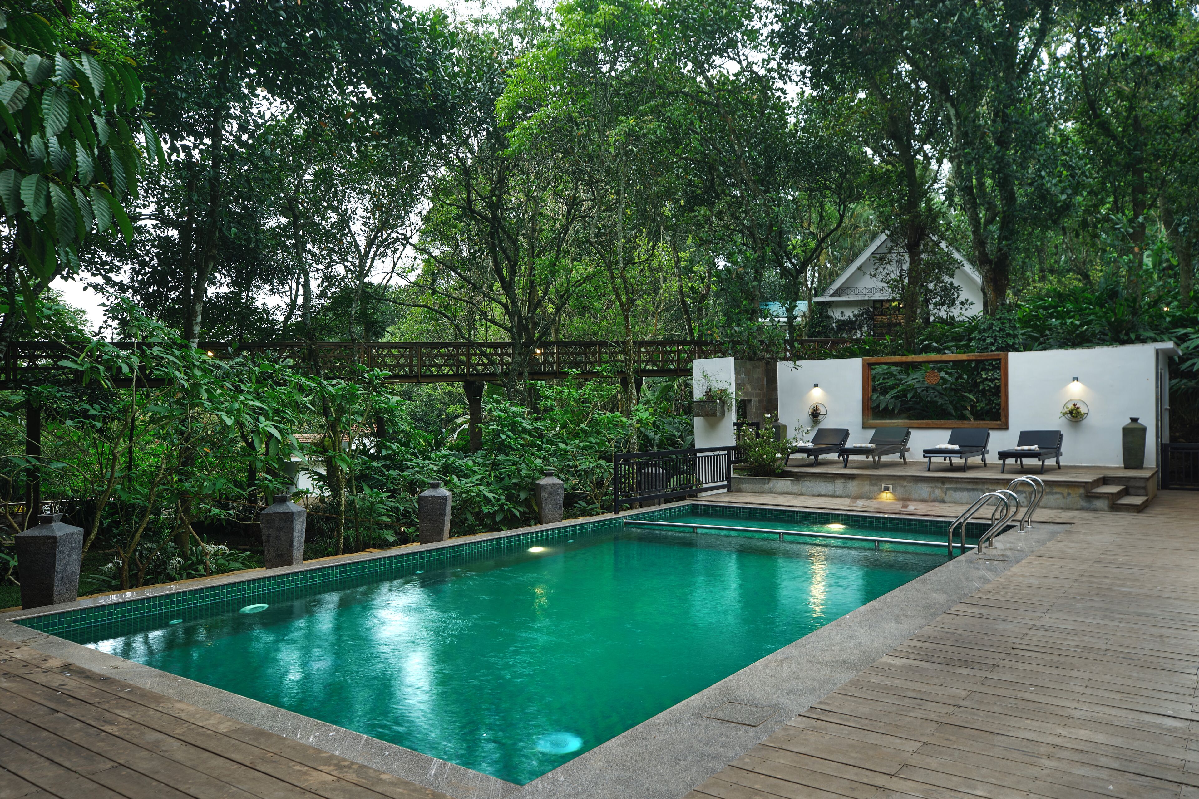A rectangular pool with a wooden deck, surrounded by lush greenery and a wooden bridge.