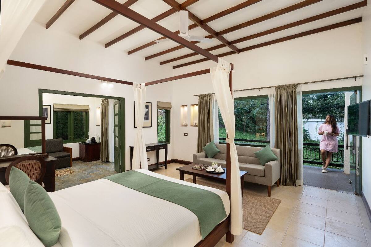 A spacious hotel room with a large bed, a sofa, a desk, and a balcony with a view of the outdoors.