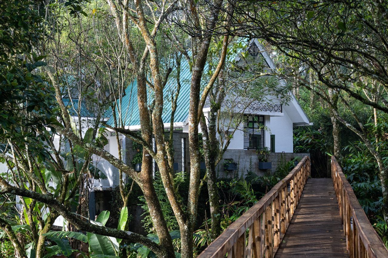 Eine Holzbrücke führt zu einem weißen Gebäude mit einem grünen Dach, umgeben von dichtem Grün.