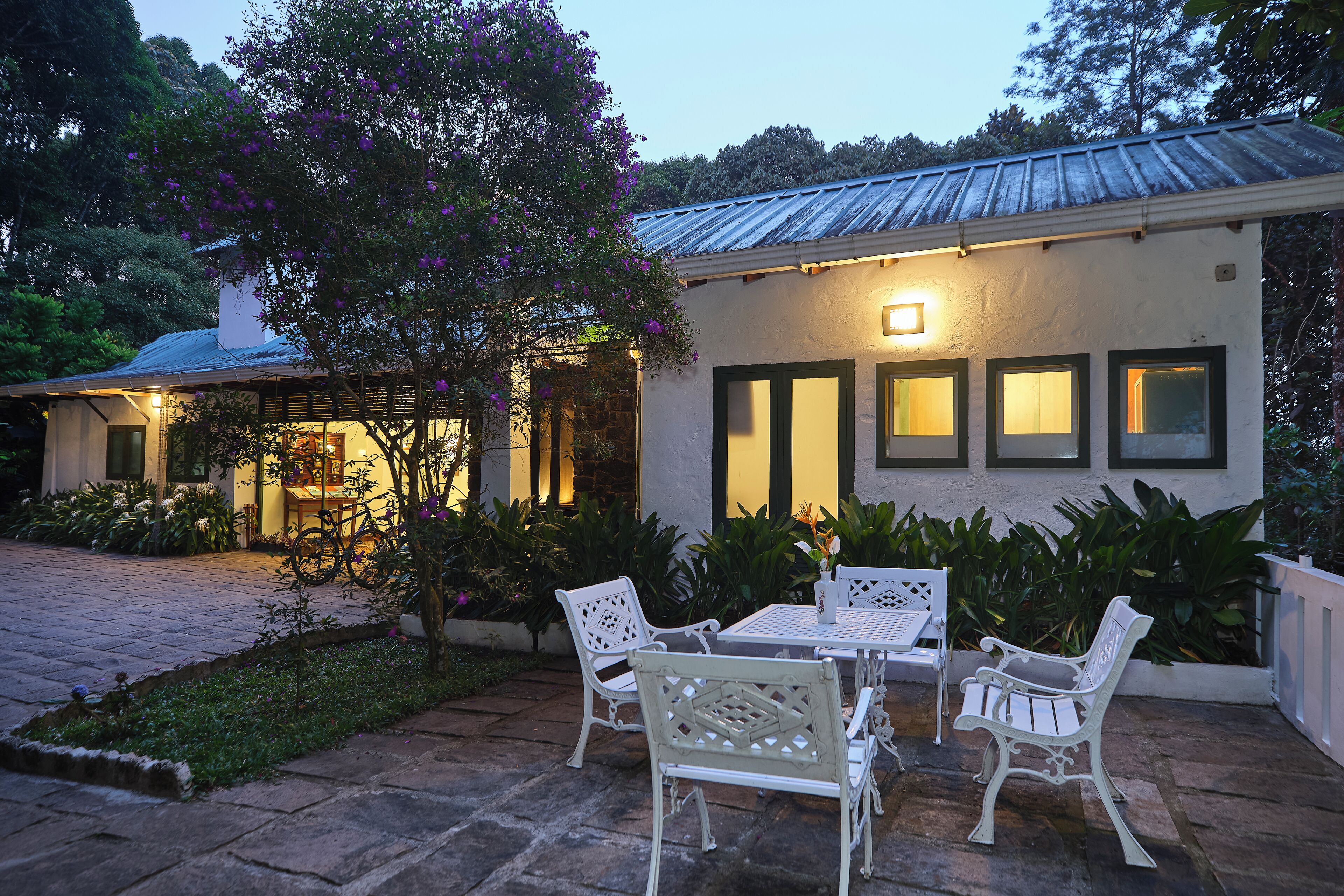 A patio with white metal chairs and a table, surrounded by lush greenery and a white building with multiple windows.