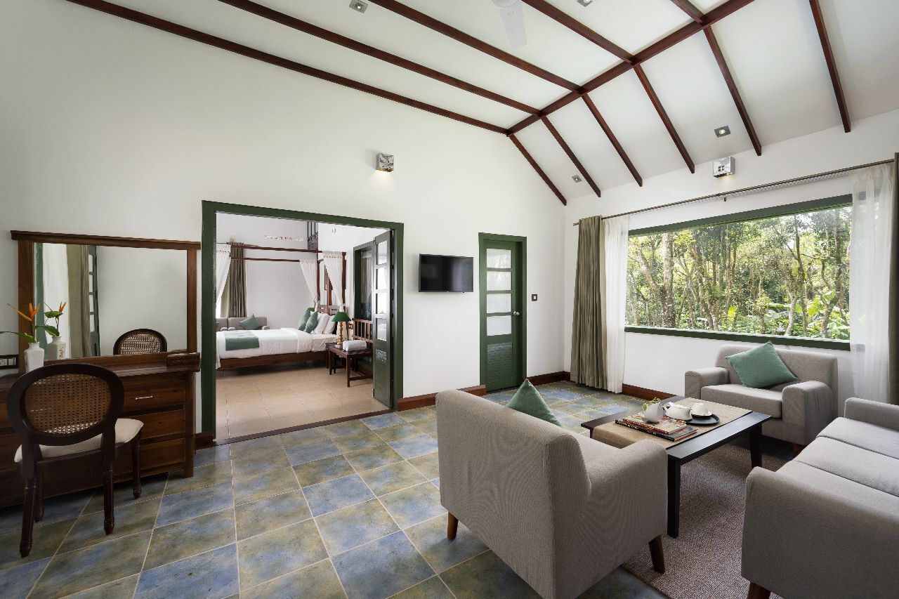 A spacious room with a bed, a desk, and a seating area. There is a large window providing a view of greenery outside.
