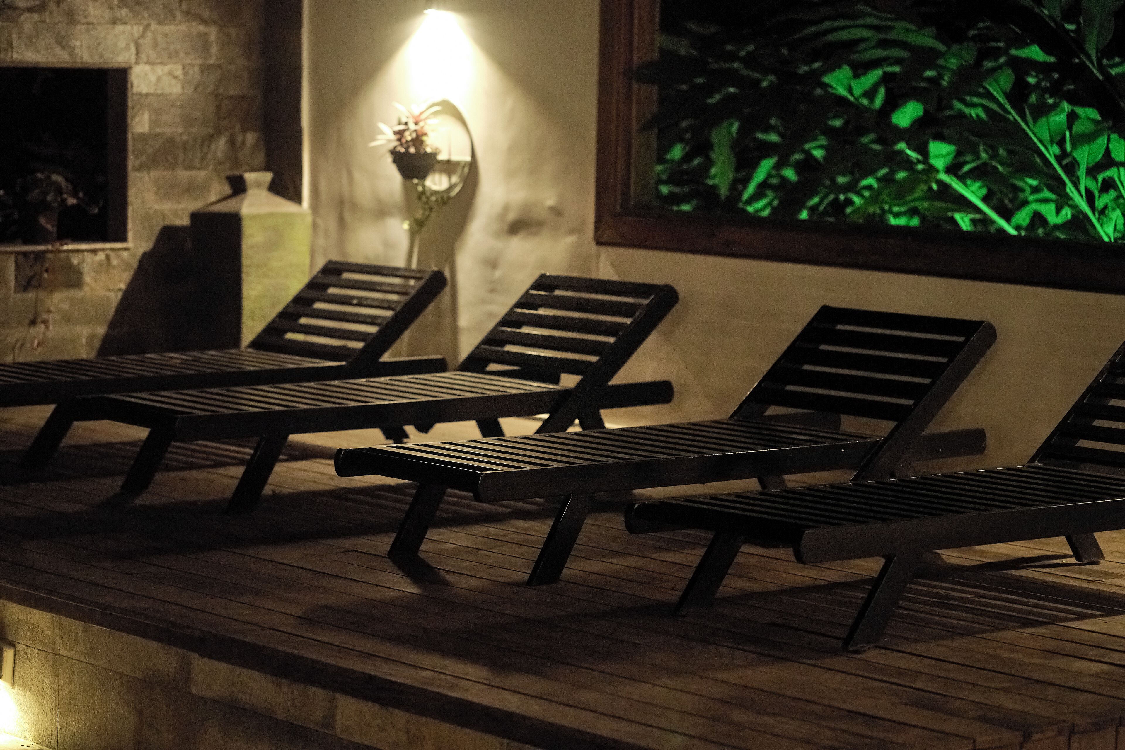 Three lounge chairs on a wooden deck with a wall-mounted light fixture and a window with a view of green foliage.