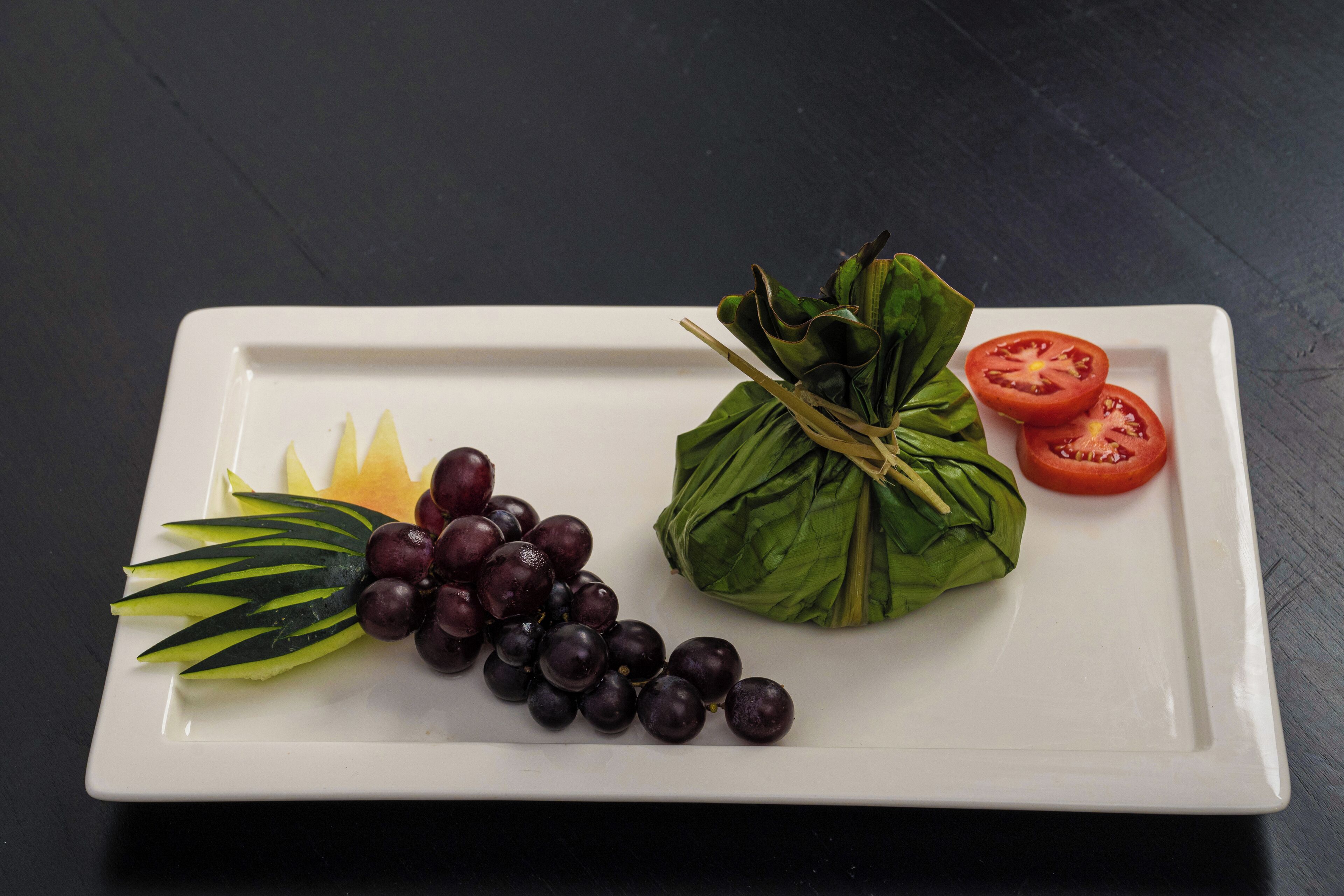 A white rectangular plate with a decorative arrangement of grapes, a wrapped item, and sliced tomatoes.