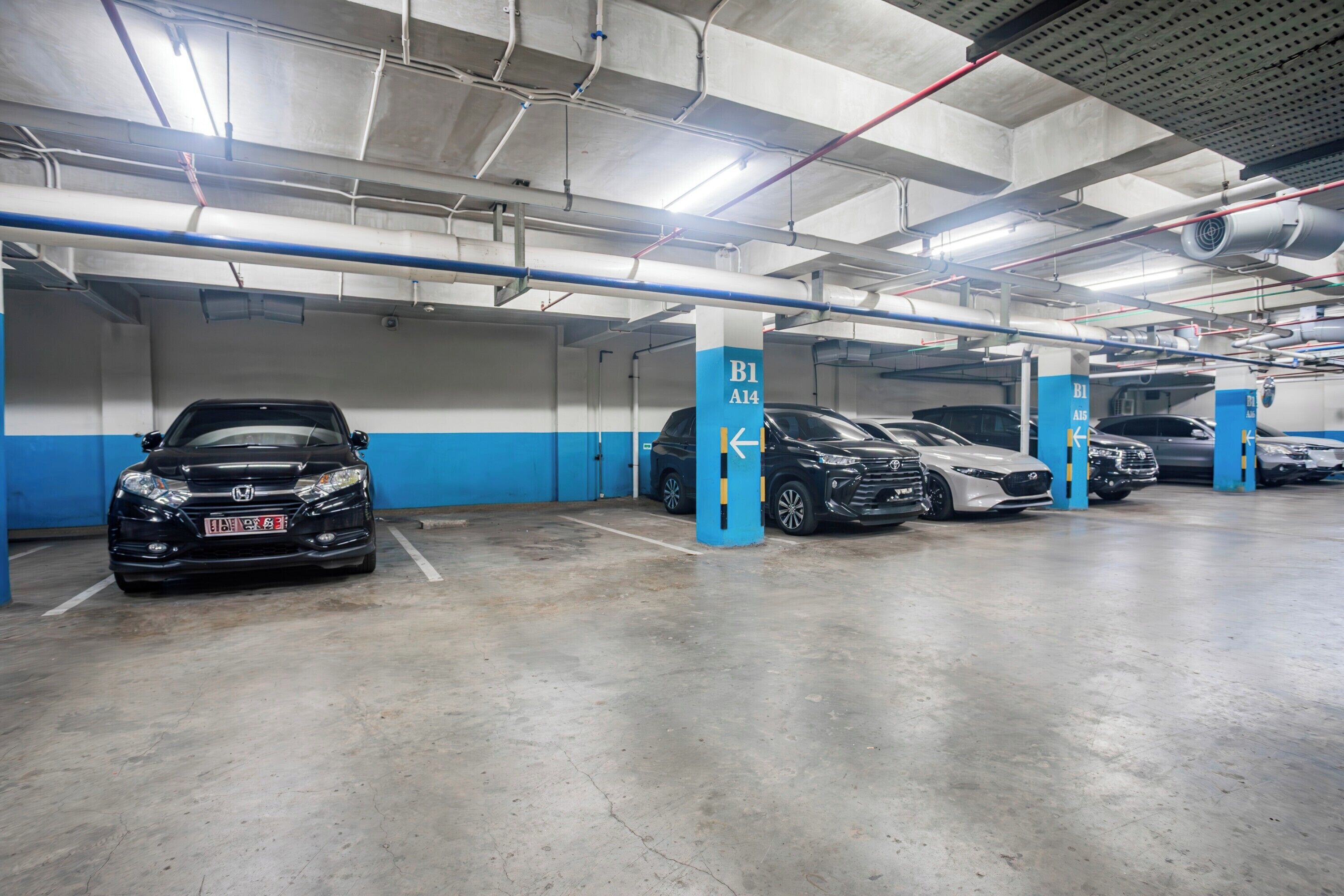 Un aparcamiento de varios niveles con coches estacionados en plazas designadas, pilares azul y blanco y tuberías del techo visibles.