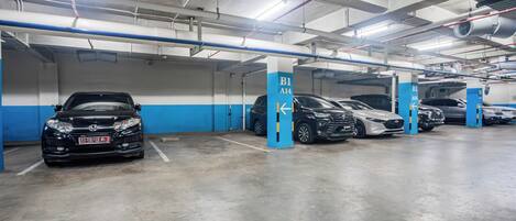 Un aparcamiento de varios niveles con coches estacionados en plazas designadas, pilares azul y blanco y tuberías del techo visibles.