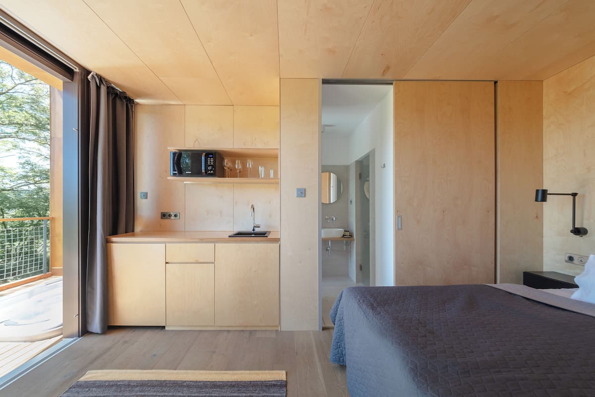 Un dormitorio moderno con techo de madera, una cama, una mesita de noche y una kitchenette integrada con microondas y copas de vino.