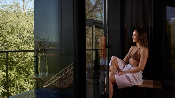 A woman sitting on a balcony with a view of trees and a building in the background. - Rock Cabins (Vigántpetend)