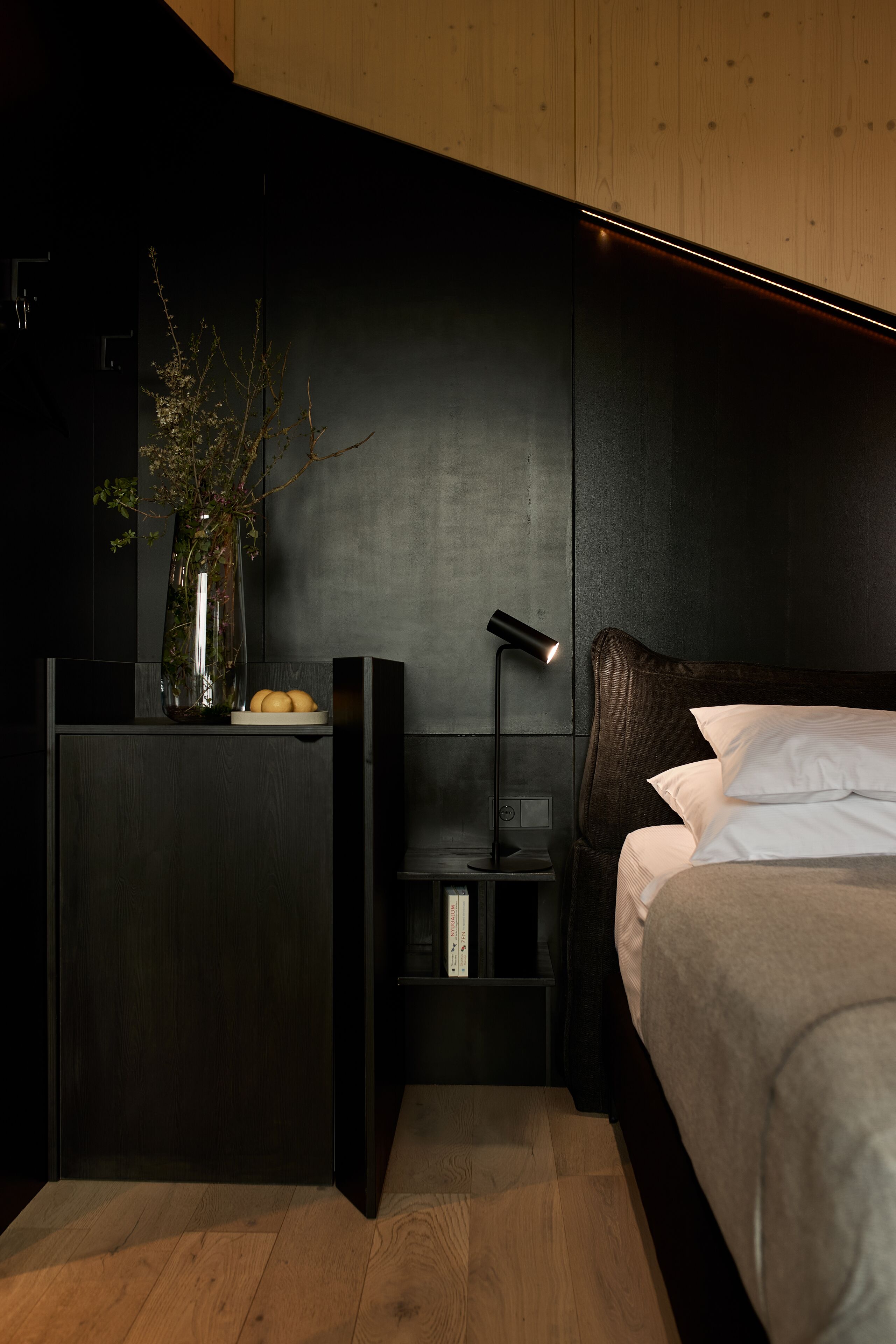 A modern bedroom with a dark wood headboard, a bedside table with a lamp, and a nightstand with a vase and books.