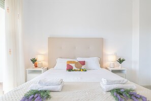 Un lit bien fait, avec du linge de lit et des serviettes blanches, deux tables de chevet avec des lampes et de petites plantes, et une tête de lit.