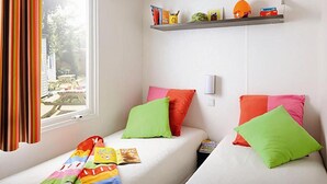 A small, bright room with two beds, colorful pillows, a window with curtains, and a shelf with various items. - Woodland Chalet in the Ardennes (Viroinval)