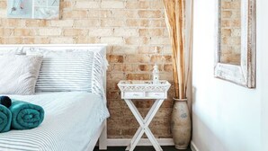 A bedroom with a brick wall, a bed with striped bedding, a small white side table, and a mirror on the wall. - Driftwood Callala Plus+ - The Perfect Jervis Bay Retreat for up to 10 guests! 🐋 (Callala Bay)