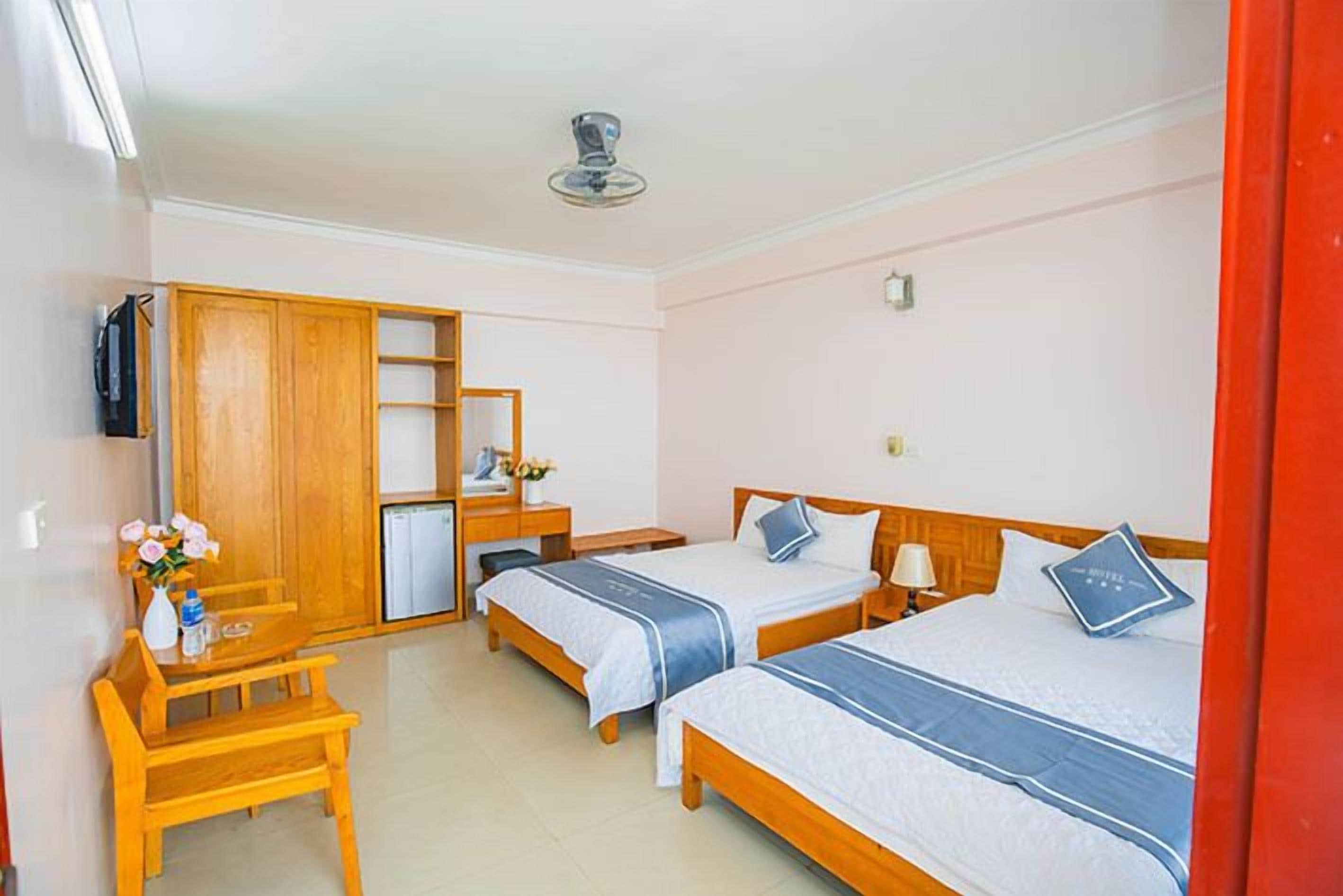 A hotel room with two single beds, a desk, a chair, a wardrobe, and a small table with a vase of flowers.