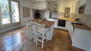 A modern kitchen with white cabinetry, a wooden island with chairs, a built-in oven, and a large window providing natural light. - Maison pic Saint Loup (Viols-le-Fort)