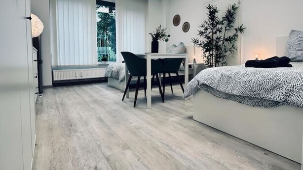 Ein modernes Schlafzimmer mit Bett, Schreibtisch und Stuhl. Ein Fenster mit Jalousien, eine Heizung und eine Pflanze sind ebenfalls vorhanden.