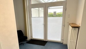 Ein Zimmer mit einer großen Schiebeglastür, die zu einem überdachten Außenbereich führt, einem Heizkörper und einem schwarzen Bürostuhl.