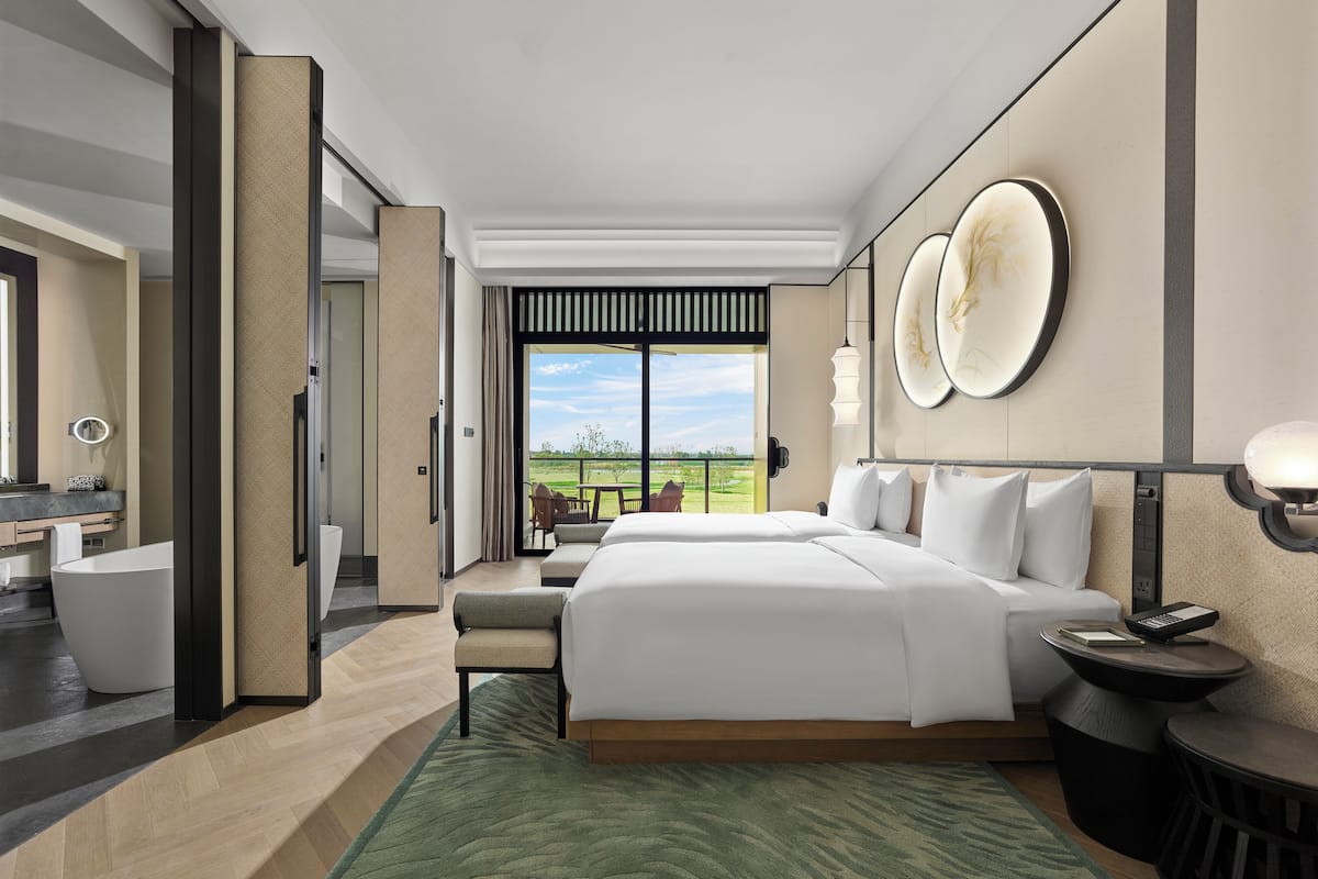 A modern hotel room with a large bed, a sitting area, a view of the outdoors, and a bathroom with a bathtub.