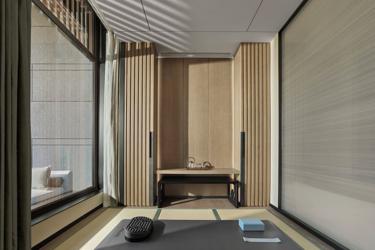 A minimalist room with tatami flooring, a tatami mat with a black object and a blue box, a low wooden table with glasses, and a sliding door.