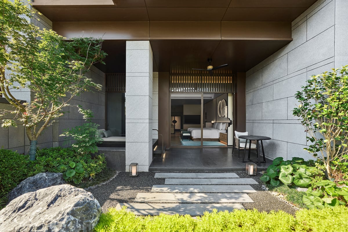 A modern hotel entrance with a covered outdoor seating area, stone walls, and a pathway leading to the entrance.