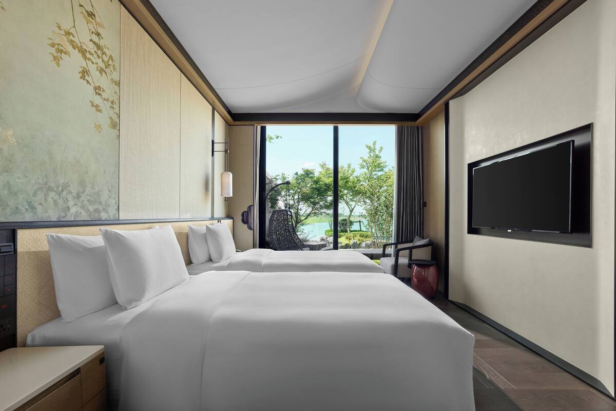 A hotel room with two beds, a large window, a flat-screen TV, and a view of greenery.